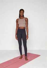 Ruda trumpa tanko tipo palaidinė ir juodos, aukštu liemeniu pasiūtos tamprės ant žmogaus, stovinčio ant rožinės jogos kilimėlio, pagamintos iš tekstūruotos medžiagos ir apta švelniai prilipusios prie kūno.