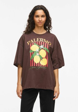 Mlada ženska z kodrastimi lasmi nosi preveliko rjavo majico z rumenimi limonami, zelenimi listi, belimi cvetovi in napisom "PALERMO".