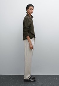 Maglione verde oliva lavorato a maglia con colletto, pantaloni crema e scarpe nere. La texture è liscia; la silhouette è snella ed allungata.