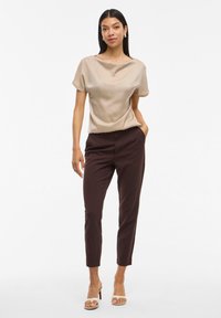 Blouse beige à coupe ample avec un col bateau, associée à un pantalon taillé marron foncé. Le modèle porte des sandales à talons ouverts.