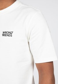 Camiseta blanca de manga corta hecha de algodón, con un logo negro "WRONG FRIENDS" impreso en el pecho y una textura suave.