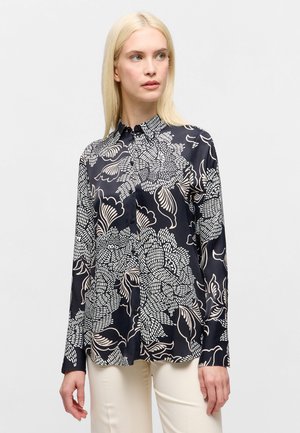 Blonde vrouw draagt een zwart overhemd met grote witte bloemmotieven en roomkleurige broek, kijkt naar links.