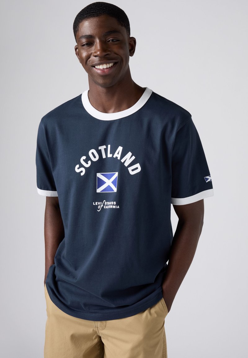 Smiling man wearing navy blue T-shirt with white trim and "Scotland" text and flag, beige pants, hands in pockets, standing against plain background.