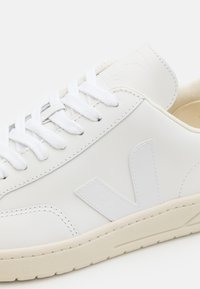 Hvit lær sneakers med hvite snører, en teksturert off-white såle, og et distinkt logo på siden. Enkel design med rene linjer.