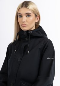 Schwarze wasserdichte Jacke mit hohem Kragen, verstellbare Kapuze, silbernen Beschlägen und Logodetails am Ärmel. Glatte Textur und taillierte Passform.