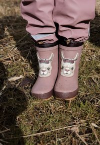 Bottes de pluie en caoutchouc rose ornées d'une illustration de lama avec une couronne de fleurs, posées sur un sol naturel avec de l'herbe et de la paille.