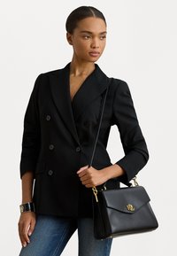Black double-breasted blazer with padded shoulders, paired with a black leather handbag featuring a gold logo and hardware, worn over blue jeans.