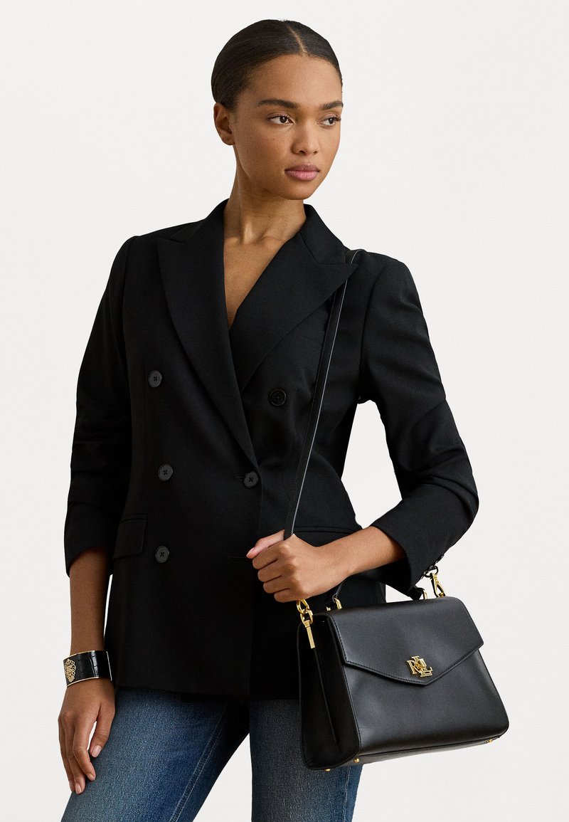 Black double-breasted blazer with padded shoulders, paired with a black leather handbag featuring a gold logo and hardware, worn over blue jeans.