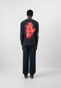 Schwarzes Langarmshirt mit einem roten Handabdruck-Motiv auf dem Rücken, kombiniert mit schwarzen Jogginghosen und grauen Sneakern. Glatte Textur.