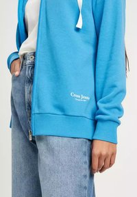 Niebieska bluza z zamkiem błyskawicznym, z białymi sznurkami i logo "Cross Jeans". Noszona na jasnoniebieskie dżinsy o luźnym kroju. Gładka tekstura.