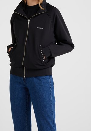 Femme portant une veste noire zippée avec le logo « THE KOOPLES » et des bordures cloutées aux poches, associée à un jean bleu, sur un fond uni.