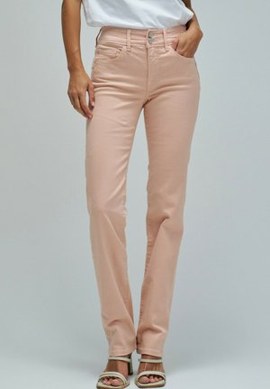 Mujer con pantalones ajustados de color rosa claro y sandalias blancas, de pie con una mano en el bolsillo frente a un fondo liso.