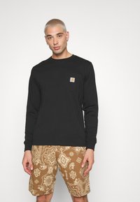 Cămașă neagră cu mânecă lungă, prevăzută cu un buzunar pe piept, având un logo galben, asortată cu pantaloni scurți cu model maro, care prezintă desene complicate.