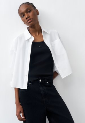 Mujer joven con una camisa blanca abierta sobre un top negro con logo, combinada con vaqueros negros de talle alto, posando contra una pared blanca.