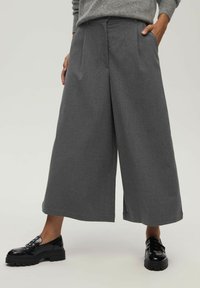 Szare spodnie typu wide-leg o dopasowanym kroju, z przodu z kantami i kieszeniami, zestawione z czarnymi lakierowanymi butami.