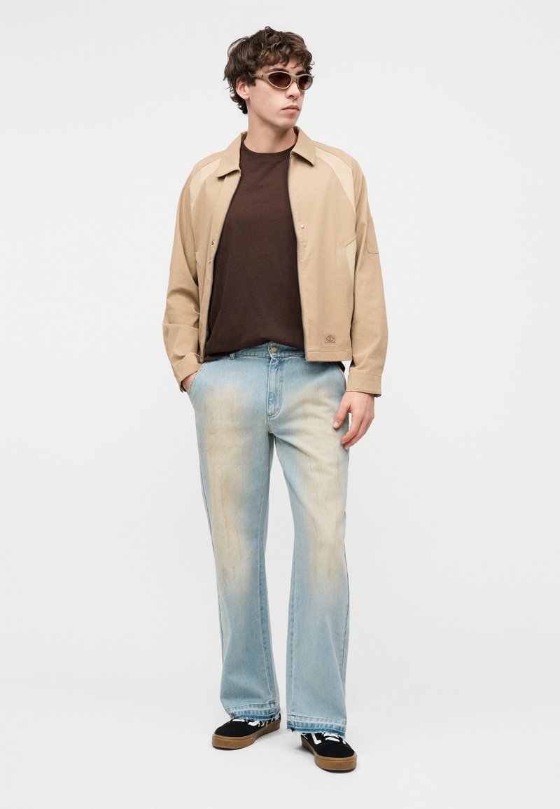 Junger Mann trägt eine beige Jacke, braunes T-Shirt, hellblaue, ausgewaschene Jeans, schwarze Sneakers und Sonnenbrille, steht mit den Händen in den Taschen.