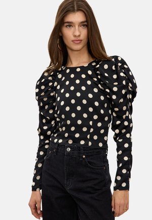 Femme portant un chemisier noir à manches bouffantes avec des pois beiges et un jean noir taille haute, posant devant un fond blanc uni.