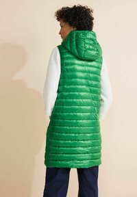 Zelená péřová vesta s kapucí, s prošívanou texturou, do pasu a se dvěma postranními kapsami. Nese se přes bílý dlouhý rukáv.