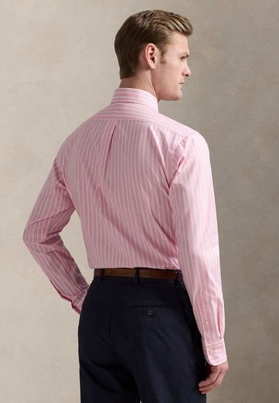 Ανοιχτό ροζ ριγέ πουκάμισο με μακριά μανίκια, μανσέτες με κουμπιά και πιέτα στην πλάτη, φορεμένο με σκούρα παντελόνια και καφέ ζώνη.