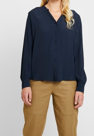 Donna che indossa una camicetta blu navy a maniche lunghe con bottoni e scollo a V, abbinata a pantaloni beige a vita alta, su uno sfondo neutro.
