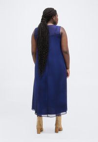 Γυναίκα με μακριά πλεξούδα, ντυμένη με αμάνικο μεσάτο φόρεμα σε ναυτικό μπλε και ταμπά μποτάκια με τακούνι, στέκεται μπροστά σε λευκό φόντο.