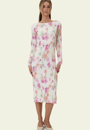 Kobieta ubrana w białą, długorękawową sukienkę midi w różowo-zielone kwiatowe wzory, w połączeniu z beżowymi sandałkami na obcasie ze zapięciem z tyłu, stojąca na jednolitym tle.