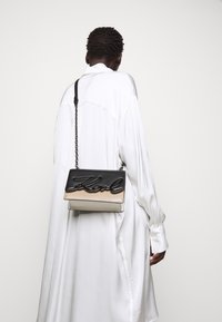 Bolso de piel negro y beige cruzado con cadena, con el logotipo "Karl" grabado en el solapa negra, textura suave.