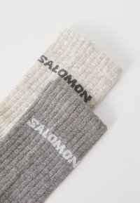 Dos pares de calcetines: uno en beige claro con una textura acanalada y el logo negro de "SALOMON"; el otro en gris oscuro con una textura similar y logo blanco.