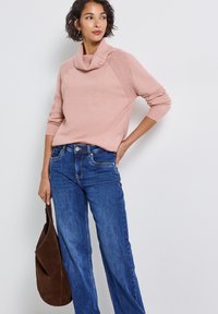 Suéter de cuello vuelto en rosa claro con acentos texturizados en los hombros, combinado con jeans de pierna ancha en azul y sosteniendo un bolso de ante marrón.