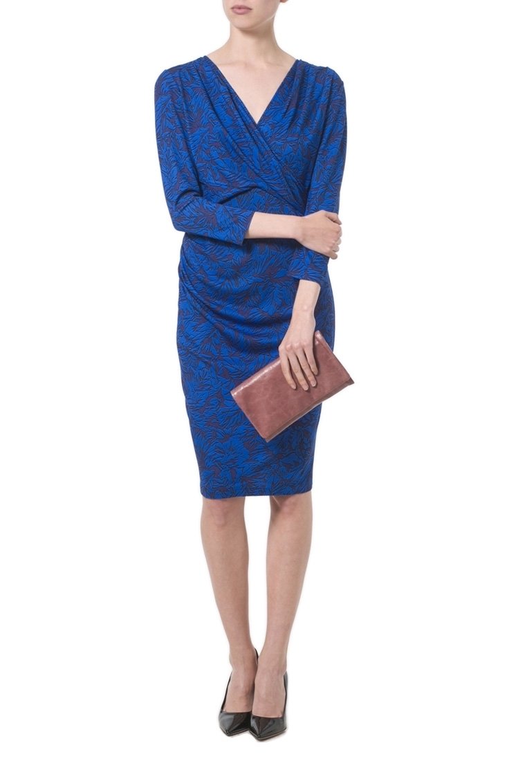 Femme portant une robe bleue à motifs longueur genou, tenant une pochette marron et portant des chaussures à talons noirs.