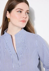 Camicia a righe blu e bianche con colletto, scollo a V e dettagli plissettati, realizzata in tessuto leggero. Accompagnata da grandi orecchini a cerchio dorati.