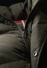 Zielona puchowa kurtka z zamkiem z przodu, prezentująca pikowany wzór, metalowy uchwyt zamka błyskawicznego oraz napy wzdłuż otwarcia.