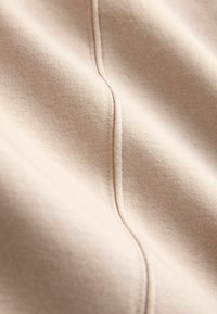 Světle béžová látka s hladkou texturou; má jemný lesk a viditelné stehy vytvářející vertikální linie. Žádné výrazné vzory ani ozdoby.