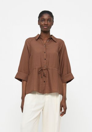 Kobieta ubrana w brązową koszulę na guziki z wiązaniem w talii i rękawami do łokcia, połączoną z białymi szerokimi spodniami.