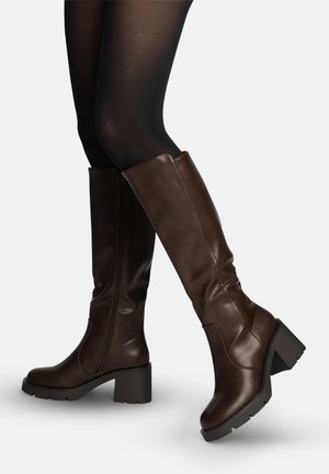 Ben i sorte strømpebukser iført mørkebrune læderstøvler op til knæet med tykke hæle og lynlåse på siden, vist mod en lys baggrund.
