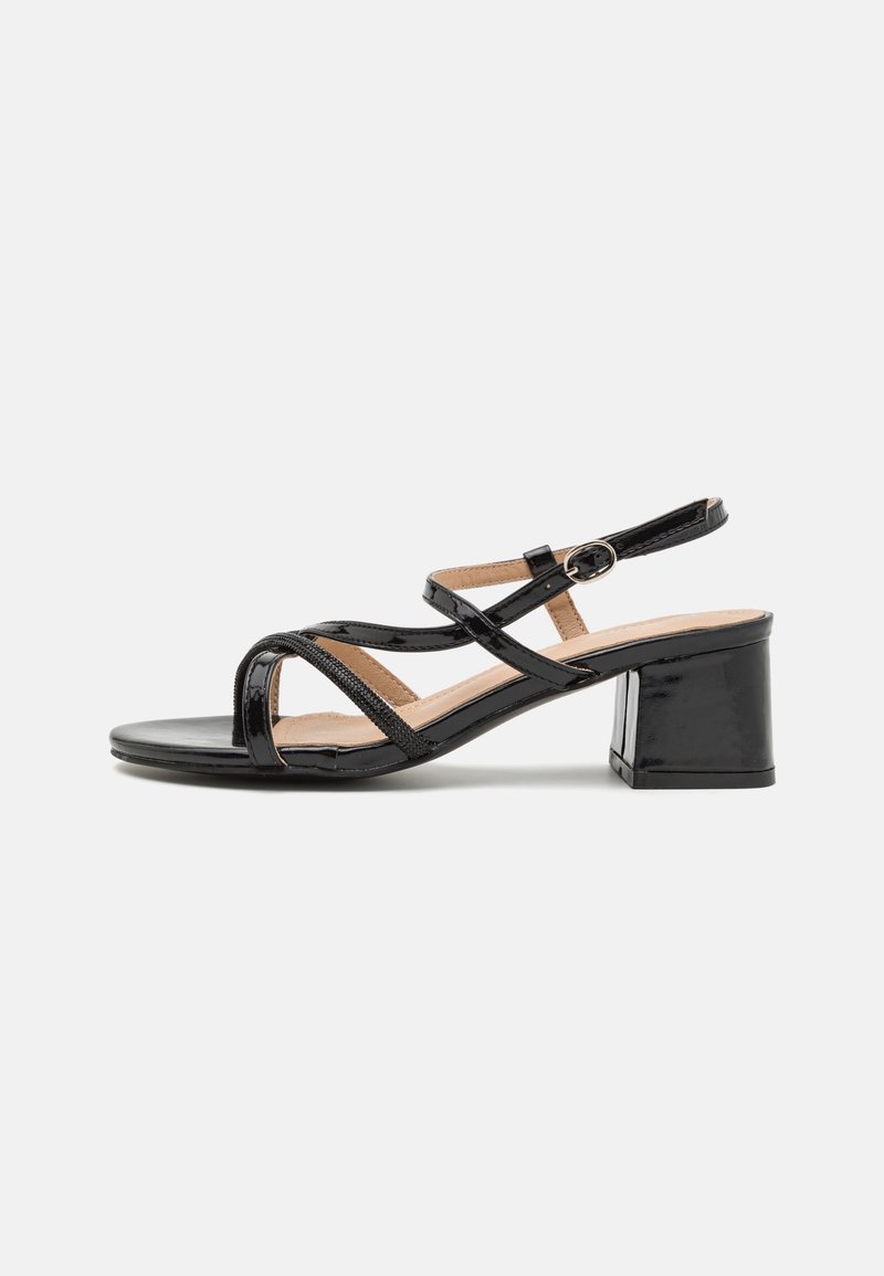 Sandale noire en cuir verni à talon bas, avec des fines brides croisées et une fermeture à boucle à la cheville sur un fond clair.