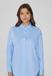 Hellblaues Hemd mit klassischem Kragen, langen Ärmeln und geradem Schnitt. Glattes Material mit leichtem Glanz. Knopfleiste vorne.