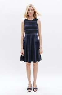 Navy-Kleid mit ärmellosem Design, das horizontale lila Streifen, einen plissierten Rock und ein tailliertes Oberteil aufweist. Kombiniert mit navy-farbenen Sandalen mit Absätzen.