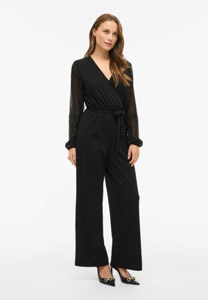 Combinaison noire avec des manches longues en voile à pois, design enveloppant, taille ceinturée, jambes larges, et associée à des talons aiguilles noirs.