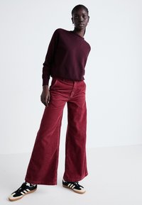 Pantaloni largi din catifea burgundy, cu buzunare frontale, asortați cu un hanorac burgundy și adidași alb-negru.