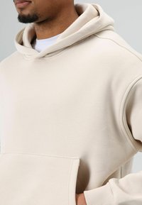 Beige hoodie van zacht stof, met een grote voorkantzak en een brede, dubbele capuchon met een ontspannen pasvorm.