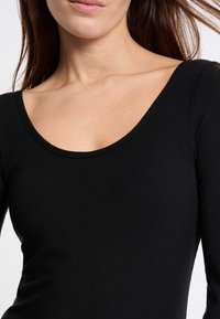 Kobieta ubrana w dopasowany czarny sweterek z długim rękawem o szerokim dekolcie, odsłaniający szyję i górną część klatki piersiowej.