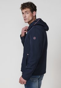 Chaqueta con capucha color azul marino de textura suave, con un acento en rojo y blanco, bolsillos laterales con cremallera y un parche de logo en la manga.