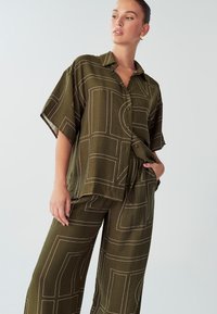 Set od maslinasto zelene košulje i hlača od lagane tkanine. Sadrži apstraktne uzorke bijelih linija i kratke rukave s kopčanjem sprijeda.