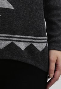 Pull gris foncé avec un motif géométrique blanc au bas, à la texture douce et à la coupe légèrement ample. Manches longues et col rond.