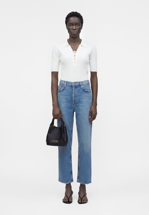 Vrouw met een wit shirt met korte mouwen, hoge blauwe jeans, zwarte sandalen met hak, houdt een kleine zwarte handtas met gouden accenten vast.