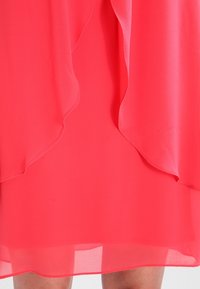 Helles Korallenkleid aus leichtem Stoff, mit fließenden, geschichteten Rüschen am Saum. Glatte Textur, am unteren Rand transparent.