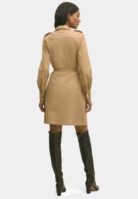 Abito beige con cintura, maniche lunghe, polsini abbottonati e spalline; indossato con stivali neri in pelle al ginocchio.