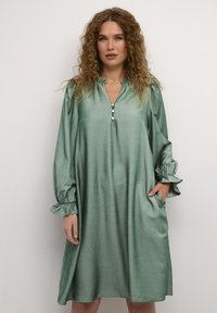 Zelena obleka do kolen, izdelana iz sijočega materiala, z V-izrezom, gumbnimi detajli in oblazinjenimi rokavi ter žepi.