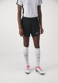 Svarte idrettsshorts med Nike-swoosh, kombinert med hvite sokker og fotballsko med rosa aksenter. Hvit og svart trøye fullfører utseendet.